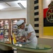 A senior military officer in white dress uniform is signing a document or guest book at a reception desk in what appears to be a Brunei Armed Forces headquarters or official military facility. The setting features the Royal Brunei Armed Forces emblem and flag prominently displayed on the wall, alongside small US and Brunei flags on the desk, indicating a bilateral military visit or diplomatic engagement. The background displays framed photographs suggesting a history gallery or ceremonial reception area.