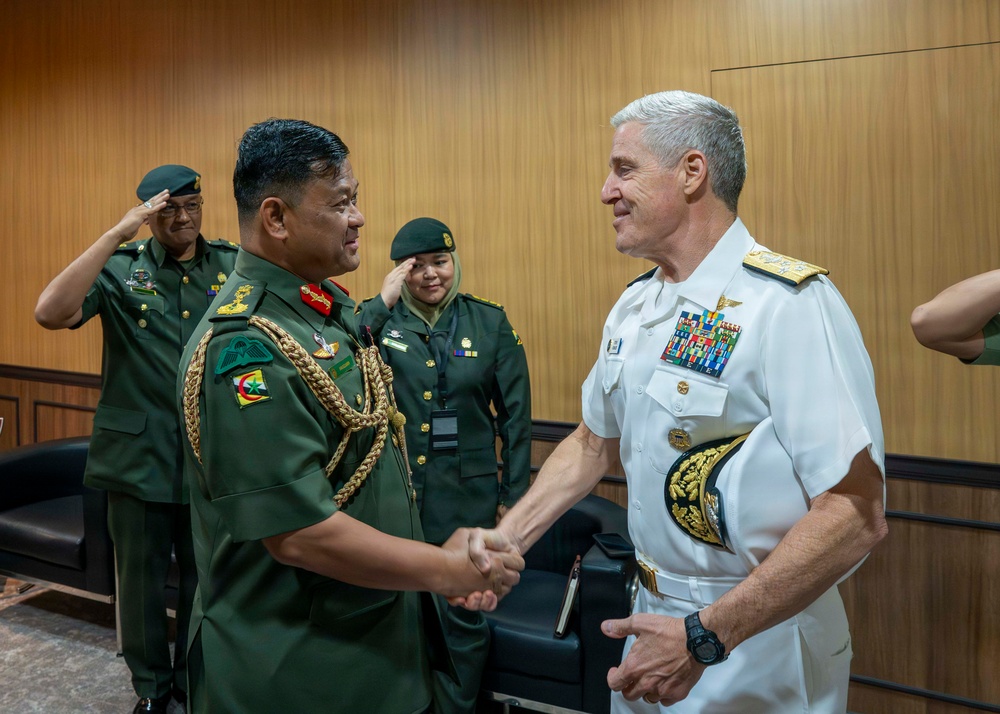 A formal military meeting is taking place between a senior officer in green army dress uniform and a senior officer in white U.S. Navy dress uniform, who are shaking hands. Several other military personnel in green uniforms are visible in the background rendering salutes. The setting appears to be an official indoor venue with wood-paneled walls.