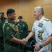A formal military meeting is taking place between a senior officer in green army dress uniform and a senior officer in white U.S. Navy dress uniform, who are shaking hands. Several other military personnel in green uniforms are visible in the background rendering salutes. The setting appears to be an official indoor venue with wood-paneled walls.