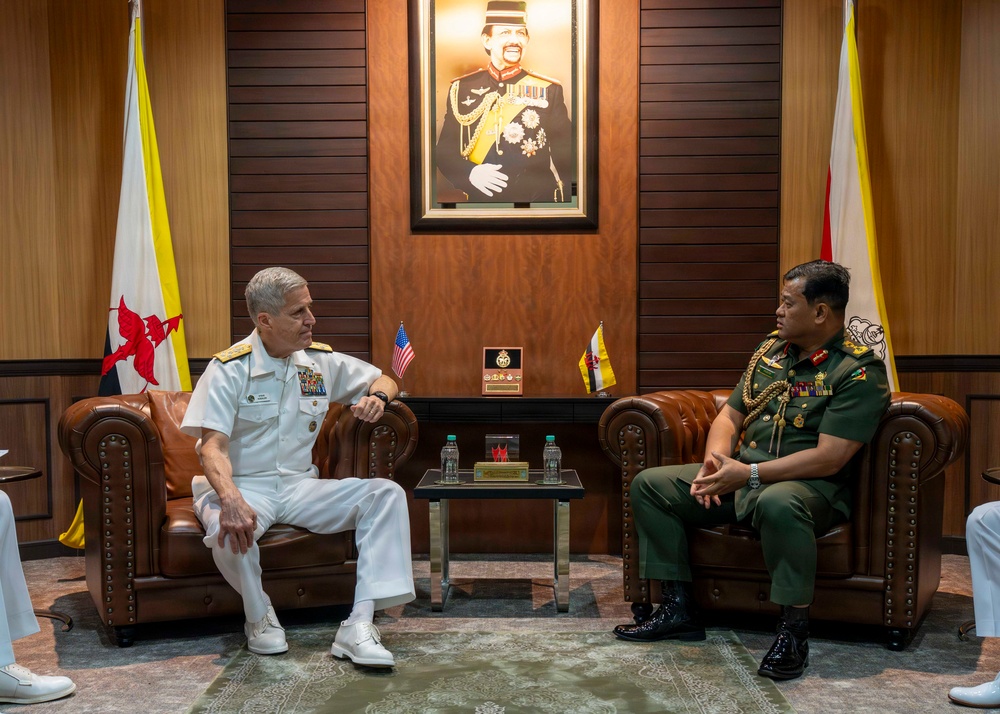 Two senior military officers are engaged in a formal bilateral meeting, seated in leather armchairs in an official reception room. One officer wears a white U.S. Navy dress uniform with admiral rank insignia, while the other wears a green military uniform with gold aiguillettes and medals, consistent with a senior Brunei Armed Forces officer. The setting includes national flags of both the United States and Brunei, along with a formal portrait on the wall.
