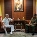 Two senior military officers are engaged in a formal bilateral meeting, seated in leather armchairs in an official reception room. One officer wears a white U.S. Navy dress uniform with admiral rank insignia, while the other wears a green military uniform with gold aiguillettes and medals, consistent with a senior Brunei Armed Forces officer. The setting includes national flags of both the United States and Brunei, along with a formal portrait on the wall.