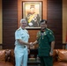 Two military officers in formal dress uniforms are shaking hands in what appears to be an official meeting room. One officer wears a white U.S. Navy dress uniform, while the other wears a dark green military uniform with medals and insignia. A framed portrait of a uniformed dignitary hangs on the wood-paneled wall behind them, flanked by two flags.