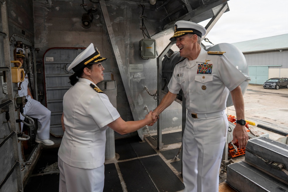 Two naval officers in white dress uniforms are shaking hands on what appears to be the deck or gangway of a naval vessel. The officer on the right displays an extensive array of service ribbons and medals, suggesting senior rank, while both wear officer's caps with gold insignia. The setting appears to be a port or naval facility.