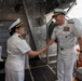 Two naval officers in white dress uniforms are shaking hands on what appears to be the deck or gangway of a naval vessel. The officer on the right displays an extensive array of service ribbons and medals, suggesting senior rank, while both wear officer's caps with gold insignia. The setting appears to be a port or naval facility.