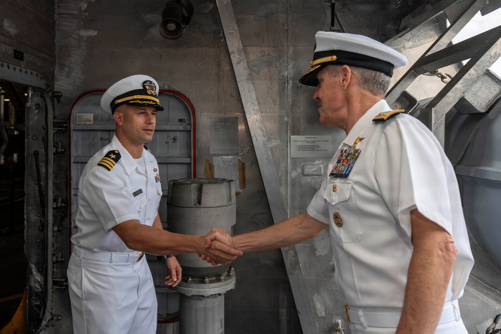 Two U.S. Navy officers in white service dress uniforms are shaking hands aboard what appears to be a naval vessel. The officer on the right displays significantly more rank insignia and ribbons, suggesting a senior flag officer meeting with a junior officer, likely a commander or lieutenant commander. The setting appears to be an interior deck or hangar bay of a warship.