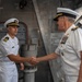 Two U.S. Navy officers in white service dress uniforms are shaking hands aboard what appears to be a naval vessel. The officer on the right displays significantly more rank insignia and ribbons, suggesting a senior flag officer meeting with a junior officer, likely a commander or lieutenant commander. The setting appears to be an interior deck or hangar bay of a warship.