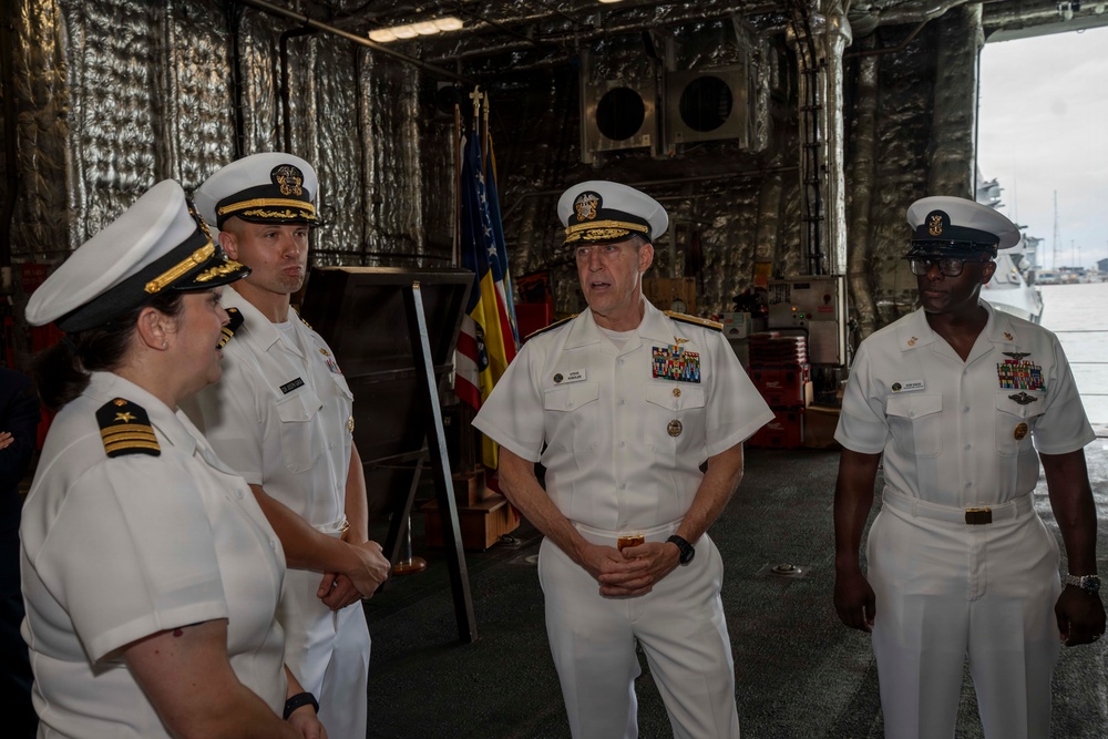 Four U.S. Navy officers in white service dress uniforms are gathered inside what appears to be a ship's hangar bay or large interior compartment. The central figure, a senior officer with extensive medal ribbons and gold-braided cap insignia consistent with flag rank (admiral), appears to be addressing or briefing the group. An American flag and what appears to be a unit or command flag are visible in the background.