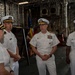 Four U.S. Navy officers in white service dress uniforms are gathered inside what appears to be a ship's hangar bay or large interior compartment. The central figure, a senior officer with extensive medal ribbons and gold-braided cap insignia consistent with flag rank (admiral), appears to be addressing or briefing the group. An American flag and what appears to be a unit or command flag are visible in the background.