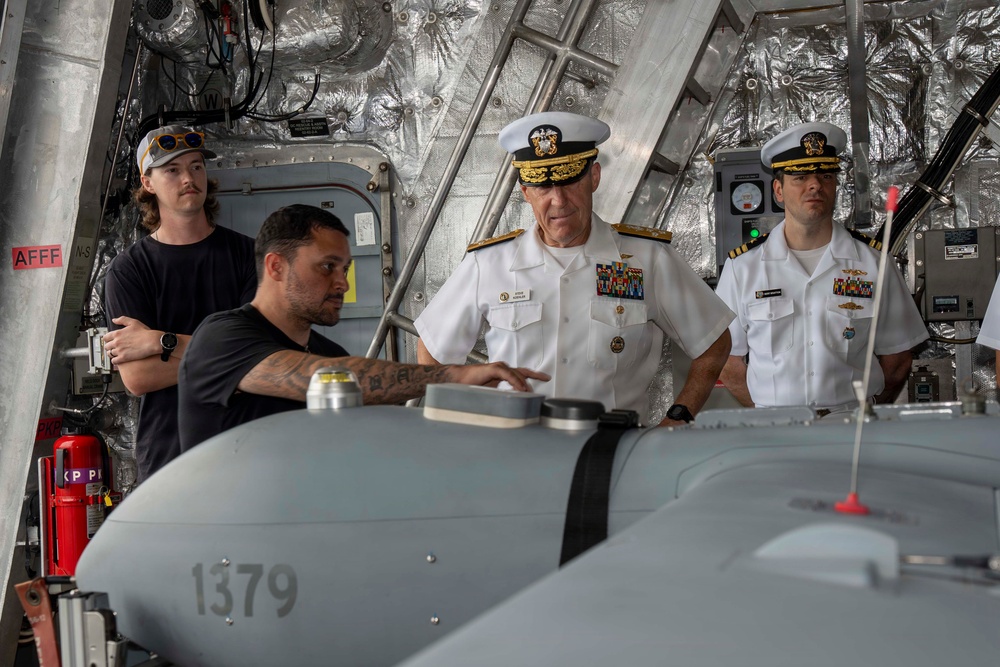 The image shows a group of four individuals inside what appears to be a military vessel or aircraft hangar bay. Two senior U.S. Navy officers in white dress uniforms with rank insignia and ribbons are observing a briefing or inspection of equipment numbered '1379', likely an unmanned aerial vehicle or torpedo. Two civilians or lower-ranking personnel in black shirts are also present, with one appearing to explain or demonstrate the equipment.