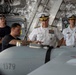 The image shows a group of four individuals inside what appears to be a military vessel or aircraft hangar bay. Two senior U.S. Navy officers in white dress uniforms with rank insignia and ribbons are observing a briefing or inspection of equipment numbered '1379', likely an unmanned aerial vehicle or torpedo. Two civilians or lower-ranking personnel in black shirts are also present, with one appearing to explain or demonstrate the equipment.