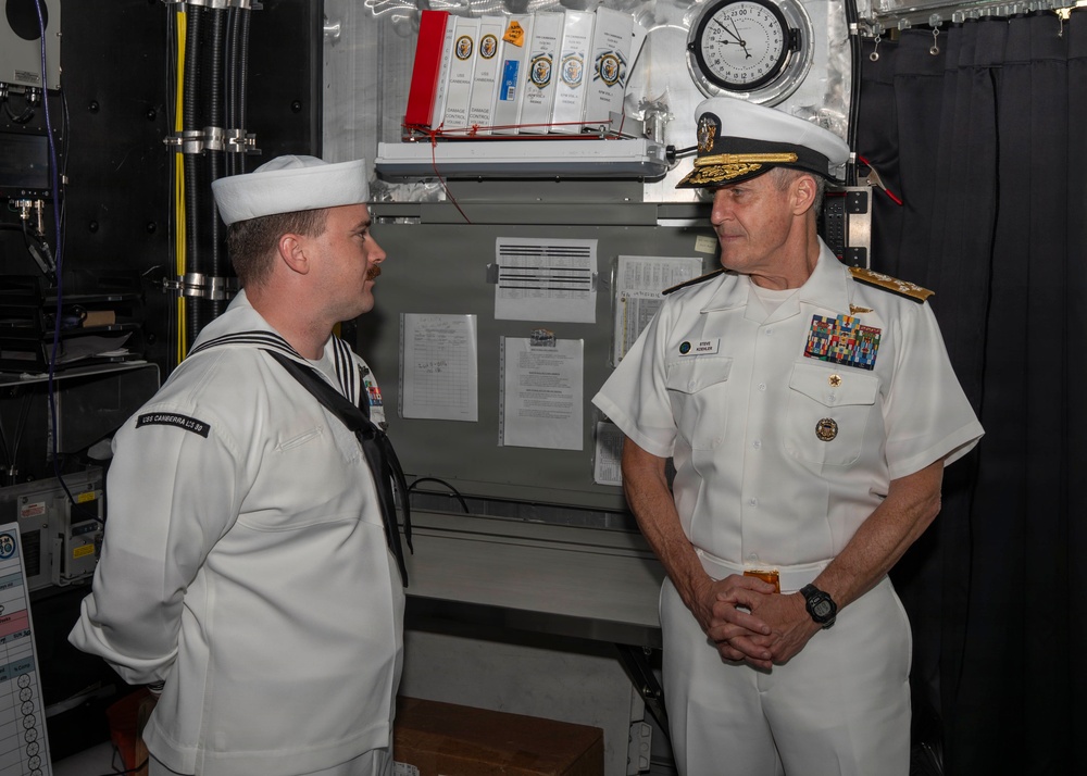 Two U.S. Navy personnel are engaged in conversation aboard what appears to be a naval vessel. On the left is an enlisted sailor in dress white uniform, and on the right is a senior flag officer (admiral) in dress white uniform with extensive ribbons and gold-braided cap. The setting appears to be an interior compartment of a ship with equipment, documents, and a clock visible in the background.