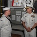 Two U.S. Navy personnel are engaged in conversation aboard what appears to be a naval vessel. On the left is an enlisted sailor in dress white uniform, and on the right is a senior flag officer (admiral) in dress white uniform with extensive ribbons and gold-braided cap. The setting appears to be an interior compartment of a ship with equipment, documents, and a clock visible in the background.