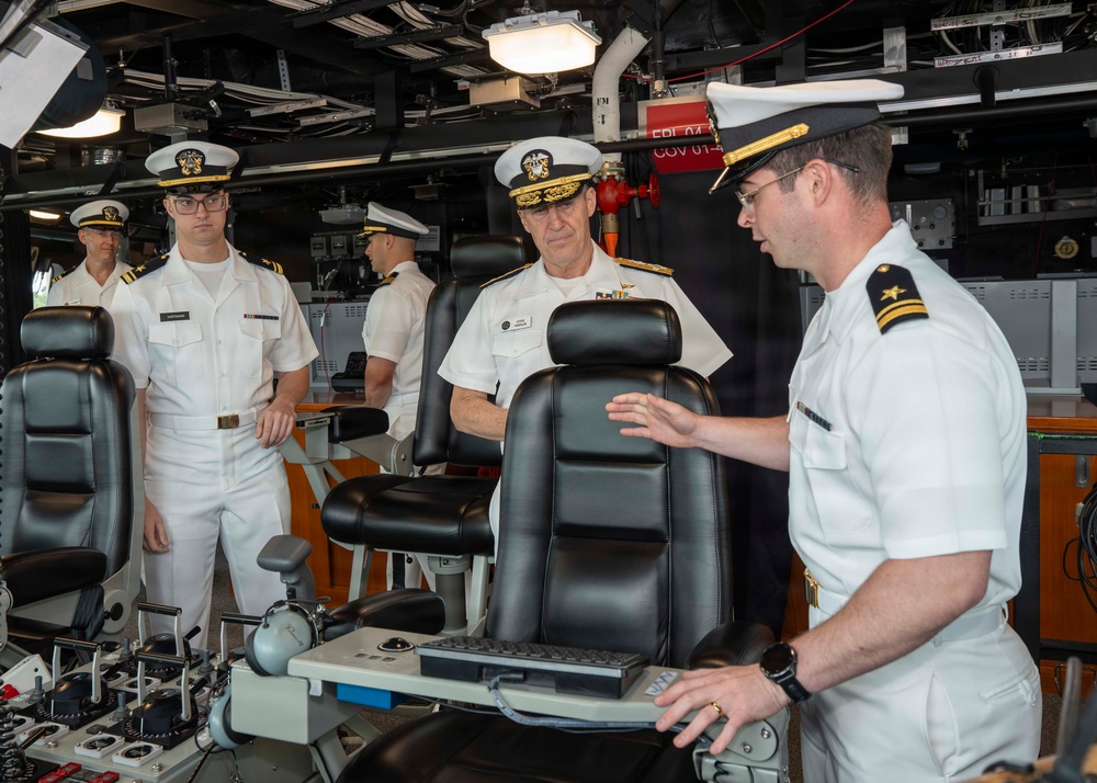 Several U.S. Navy personnel in white service uniforms are gathered on what appears to be the bridge or combat information center of a naval vessel. A senior officer with admiral-rank insignia (multiple stars on shoulder boards) is receiving a briefing or tour from a junior officer at a control console. Additional officers and enlisted personnel are present in the background observing.