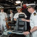 Several U.S. Navy personnel in white service uniforms are gathered on what appears to be the bridge or combat information center of a naval vessel. A senior officer with admiral-rank insignia (multiple stars on shoulder boards) is receiving a briefing or tour from a junior officer at a control console. Additional officers and enlisted personnel are present in the background observing.