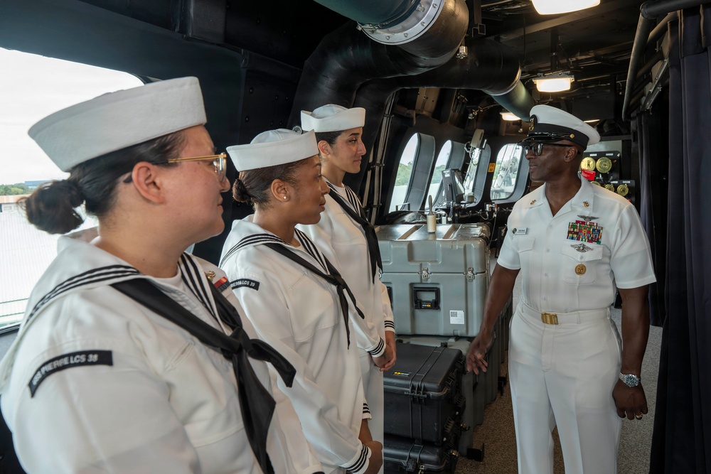 A senior U.S. Navy officer in dress white uniform is addressing three junior enlisted sailors (also in dress whites with Dixie cup covers) aboard what appears to be the bridge or interior passageway of a naval vessel. The officer displays multiple service ribbons and warfare insignia, indicating significant experience and rank. The setting appears to be an official visit, briefing, or orientation aboard a Littoral Combat Ship (LCS), as suggested by the name tags visible on the sailors' uniforms.