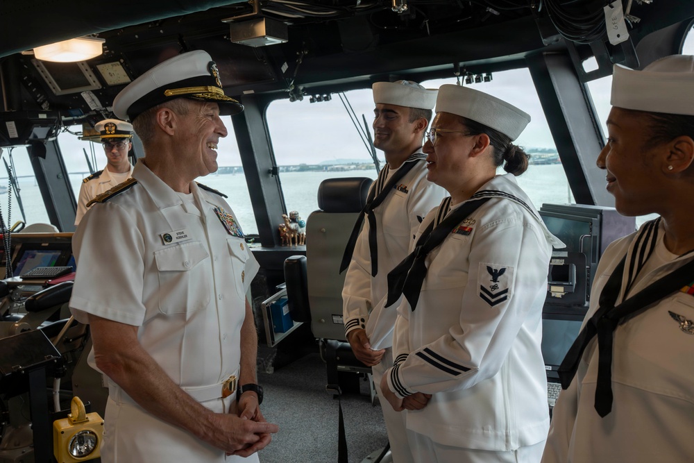 A senior U.S. Navy flag officer in white service dress uniform is engaged in a friendly conversation with enlisted sailors aboard what appears to be a ship's bridge or pilothouse. The officer, wearing a combination cover with gold insignia and multiple ribbons/medals, is smiling and interacting with at least three sailors in white crackerjack uniforms. The setting is clearly a naval vessel's command area, with navigation equipment and water visible through the windows.