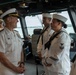A senior U.S. Navy flag officer in white service dress uniform is engaged in a friendly conversation with enlisted sailors aboard what appears to be a ship's bridge or pilothouse. The officer, wearing a combination cover with gold insignia and multiple ribbons/medals, is smiling and interacting with at least three sailors in white crackerjack uniforms. The setting is clearly a naval vessel's command area, with navigation equipment and water visible through the windows.