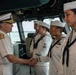 A senior U.S. Navy flag officer in white service dress uniform is shaking hands with enlisted sailors aboard what appears to be a naval vessel's bridge or pilothouse. The officer, distinguished by gold sleeve stripes and a combination cover with gold insignia, is greeting a line of female sailors in white crackerjack uniforms. The setting appears to be an official visit or inspection aboard a commissioned U.S. Navy ship.