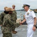 A senior naval officer in white dress uniform is shaking hands with a service member in digital camouflage (NWU Type III) aboard what appears to be a naval vessel at a port. Two additional personnel wearing U.S. Coast Guard caps and camouflage uniforms stand in the background observing the interaction. The setting appears to be an official visit or inspection at a waterfront military installation.