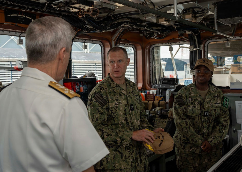 Three military personnel are engaged in conversation aboard what appears to be a ship's bridge or command center. Two individuals in U.S. Navy/Coast Guard digital camouflage uniforms (name tapes visible: 'ROSS' and 'GILLIAM') are speaking with a senior officer in a white dress uniform with gold shoulder boards. One of the camouflage-uniformed personnel is holding what appears to be a Coast Guard cover/cap, suggesting a ceremonial exchange or presentation.