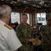 Three military personnel are engaged in conversation aboard what appears to be a ship's bridge or command center. Two individuals in U.S. Navy/Coast Guard digital camouflage uniforms (name tapes visible: 'ROSS' and 'GILLIAM') are speaking with a senior officer in a white dress uniform with gold shoulder boards. One of the camouflage-uniformed personnel is holding what appears to be a Coast Guard cover/cap, suggesting a ceremonial exchange or presentation.
