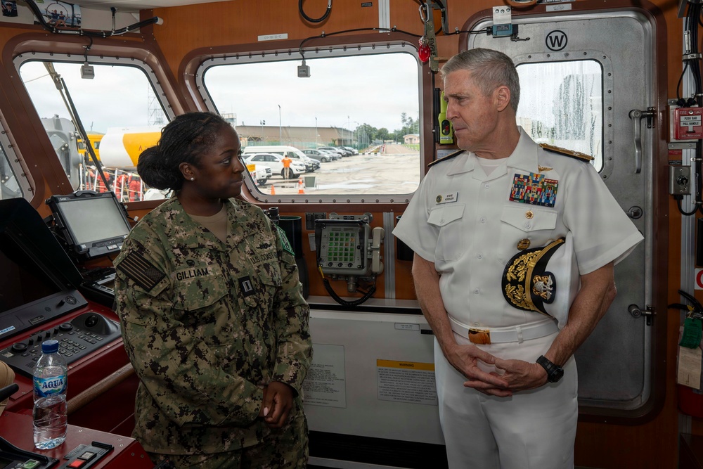 Two U.S. military personnel are engaged in conversation aboard what appears to be a vessel's bridge or command area. One is a junior enlisted/officer in Navy Working Uniform (NWU) camouflage with the name tape 'GILLIAM' and a captain's rank insignia, while the other is a senior flag officer in white dress uniform adorned with numerous ribbons and gold shoulder boards indicating admiral rank. The setting appears to be a ship's bridge with navigation equipment and windows overlooking a port facility.