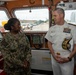 Two U.S. military personnel are engaged in conversation aboard what appears to be a vessel's bridge or command area. One is a junior enlisted/officer in Navy Working Uniform (NWU) camouflage with the name tape 'GILLIAM' and a captain's rank insignia, while the other is a senior flag officer in white dress uniform adorned with numerous ribbons and gold shoulder boards indicating admiral rank. The setting appears to be a ship's bridge with navigation equipment and windows overlooking a port facility.