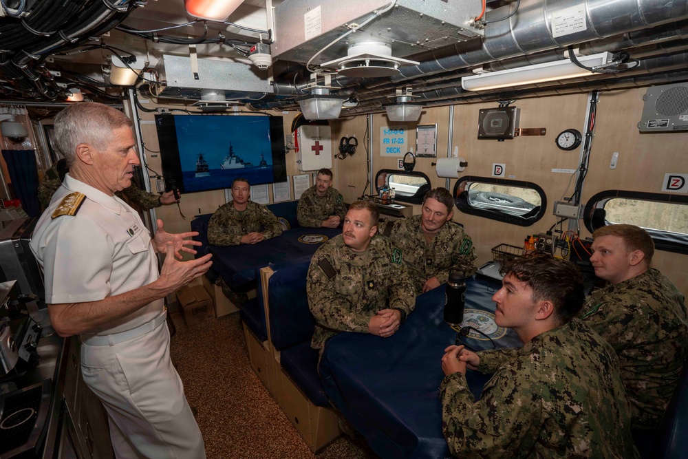 A senior naval officer in white dress uniform addresses a group of enlisted sailors wearing Navy Working Uniform (NWU) camouflage inside what appears to be a shipboard berthing or briefing compartment. The officer, displaying flag-rank insignia (admiral-level), is gesturing while speaking to approximately six sailors seated around a blue-covered surface. A monitor in the background displays naval vessels at sea.