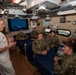 A senior naval officer in white dress uniform addresses a group of enlisted sailors wearing Navy Working Uniform (NWU) camouflage inside what appears to be a shipboard berthing or briefing compartment. The officer, displaying flag-rank insignia (admiral-level), is gesturing while speaking to approximately six sailors seated around a blue-covered surface. A monitor in the background displays naval vessels at sea.