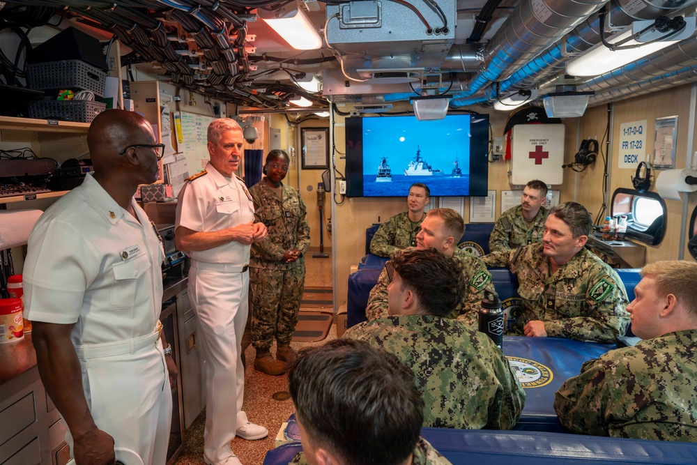 Two senior naval officers in white dress uniforms are addressing a group of enlisted and junior personnel in camouflage uniforms inside what appears to be a naval vessel's interior compartment. The setting appears to be a briefing or command visit, with personnel seated and standing in a confined shipboard space. A monitor in the background displays naval vessels at sea.