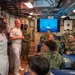 Two senior naval officers in white dress uniforms are addressing a group of enlisted and junior personnel in camouflage uniforms inside what appears to be a naval vessel's interior compartment. The setting appears to be a briefing or command visit, with personnel seated and standing in a confined shipboard space. A monitor in the background displays naval vessels at sea.