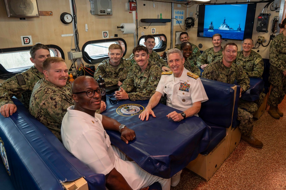 A group of U.S. Navy personnel are gathered around a table in what appears to be a mess or common area aboard a naval vessel. A senior officer in white dress uniform, displaying multiple ribbons and admiral-rank insignia, is seated at the center alongside enlisted sailors and junior officers in camouflage working uniforms. A naval ship is visible on a monitor in the background.