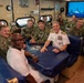 A group of U.S. Navy personnel are gathered around a table in what appears to be a mess or common area aboard a naval vessel. A senior officer in white dress uniform, displaying multiple ribbons and admiral-rank insignia, is seated at the center alongside enlisted sailors and junior officers in camouflage working uniforms. A naval ship is visible on a monitor in the background.