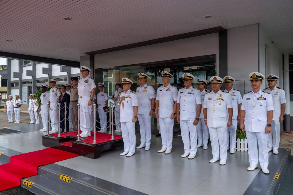 A formal military ceremony is taking place at what appears to be a naval headquarters or official building. A group of naval officers in white dress uniforms are assembled in formation, with two senior officers standing on a raised red-carpeted dais. The gathering appears to be a multinational event, with officers of varying nationalities and one taller Western officer visible among predominantly Asian naval personnel.