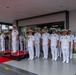 A formal military ceremony is taking place at what appears to be a naval headquarters or official building. A group of naval officers in white dress uniforms are assembled in formation, with two senior officers standing on a raised red-carpeted dais. The gathering appears to be a multinational event, with officers of varying nationalities and one taller Western officer visible among predominantly Asian naval personnel.
