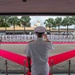 A formal military naval ceremony is taking place on a parade ground, with a senior officer in white dress uniform saluting a guard of honor lined up in formation. Red carpets are arranged in a V-shape or star pattern on the tarmac, indicating a high-protocol ceremonial reception. Two flags are visible in the background near a low-rise building surrounded by palm trees, suggesting a tropical location consistent with Southeast Asia.