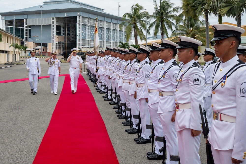 A formal military honor guard ceremony is taking place outdoors at what appears to be a naval base. Senior naval officers in dress uniforms are walking along a red carpet while a line of uniformed honor guard personnel stand at attention. The setting includes a large industrial or hangar-style building and palm trees in the background, suggesting a tropical location.