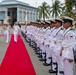 A formal military honor guard ceremony is taking place outdoors at what appears to be a naval base. Senior naval officers in dress uniforms are walking along a red carpet while a line of uniformed honor guard personnel stand at attention. The setting includes a large industrial or hangar-style building and palm trees in the background, suggesting a tropical location.
