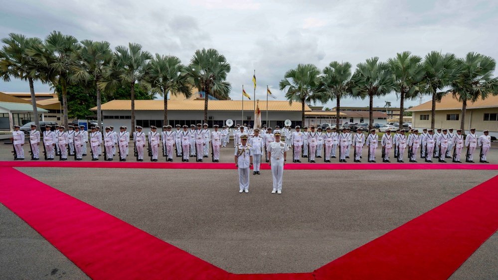 A formal military parade or ceremonial event is taking place on a paved parade ground, with two senior officers standing at the center on a red carpet arrangement in a V or arrow shape. A large formation of naval personnel dressed in white uniforms are standing at attention in rows behind them. The setting appears to be a tropical military base, indicated by palm trees and low-rise buildings in the background.