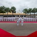A formal military parade or ceremonial event is taking place on a paved parade ground, with two senior officers standing at the center on a red carpet arrangement in a V or arrow shape. A large formation of naval personnel dressed in white uniforms are standing at attention in rows behind them. The setting appears to be a tropical military base, indicated by palm trees and low-rise buildings in the background.