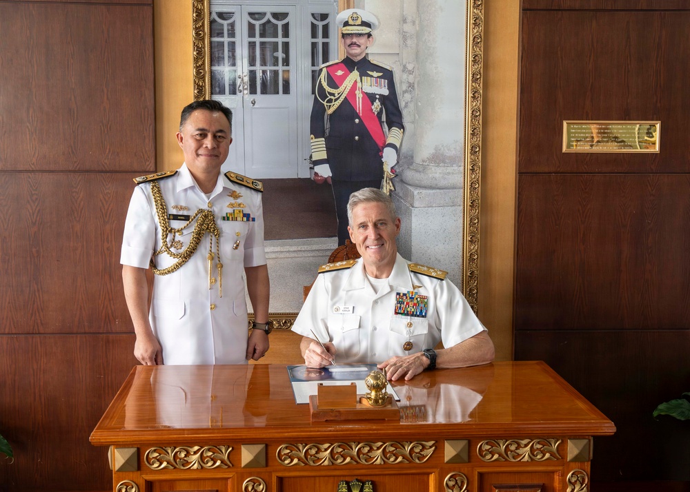 Two naval officers in white dress uniforms are posed at an ornate wooden desk, with one seated and signing a document while the other stands beside him. Behind them hangs a large formal portrait of a senior military official in ceremonial dress uniform with a red sash. The setting appears to be an official military office, likely during a formal diplomatic or ceremonial visit.