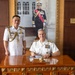 Two naval officers in white dress uniforms are posed at an ornate wooden desk, with one seated and signing a document while the other stands beside him. Behind them hangs a large formal portrait of a senior military official in ceremonial dress uniform with a red sash. The setting appears to be an official military office, likely during a formal diplomatic or ceremonial visit.