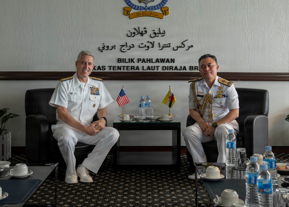Two senior naval officers are seated in a formal meeting room at the Royal Brunei Navy headquarters (Markas Tentera Laut Diraja Brunei), as indicated by the signage on the wall reading 'Bilik Pahlawan' and 'Markas Tentera Laut Diraja Brunei'. One officer wears a US Navy white uniform with ribbons and insignia, while the other wears a Royal Brunei Navy white dress uniform adorned with ceremonial gold aiguillettes and medals. Small US and Brunei national flags are displayed on a table between them, indicating a bilateral military diplomatic meeting.