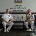 Two senior naval officers are seated in a formal meeting room at the Royal Brunei Navy headquarters (Markas Tentera Laut Diraja Brunei), as indicated by the signage on the wall reading 'Bilik Pahlawan' and 'Markas Tentera Laut Diraja Brunei'. One officer wears a US Navy white uniform with ribbons and insignia, while the other wears a Royal Brunei Navy white dress uniform adorned with ceremonial gold aiguillettes and medals. Small US and Brunei national flags are displayed on a table between them, indicating a bilateral military diplomatic meeting.