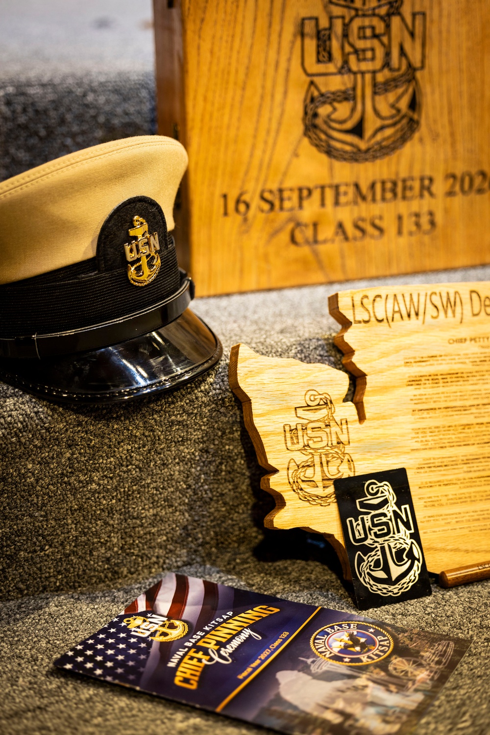 The image depicts a collection of U.S. Navy Chief Petty Officer pinning ceremony memorabilia arranged on a carpeted surface. Items include a Chief Petty Officer combination cover (hat), engraved wooden plaques and keepsakes bearing the USN anchor insignia, and a program booklet from Naval Base Kitsap. The wooden box in the background is engraved with '16 September 202_ Class 133,' indicating this is from a Chief Petty Officer pinning ceremony.