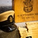 The image depicts a collection of U.S. Navy Chief Petty Officer pinning ceremony memorabilia arranged on a carpeted surface. Items include a Chief Petty Officer combination cover (hat), engraved wooden plaques and keepsakes bearing the USN anchor insignia, and a program booklet from Naval Base Kitsap. The wooden box in the background is engraved with '16 September 202_ Class 133,' indicating this is from a Chief Petty Officer pinning ceremony.
