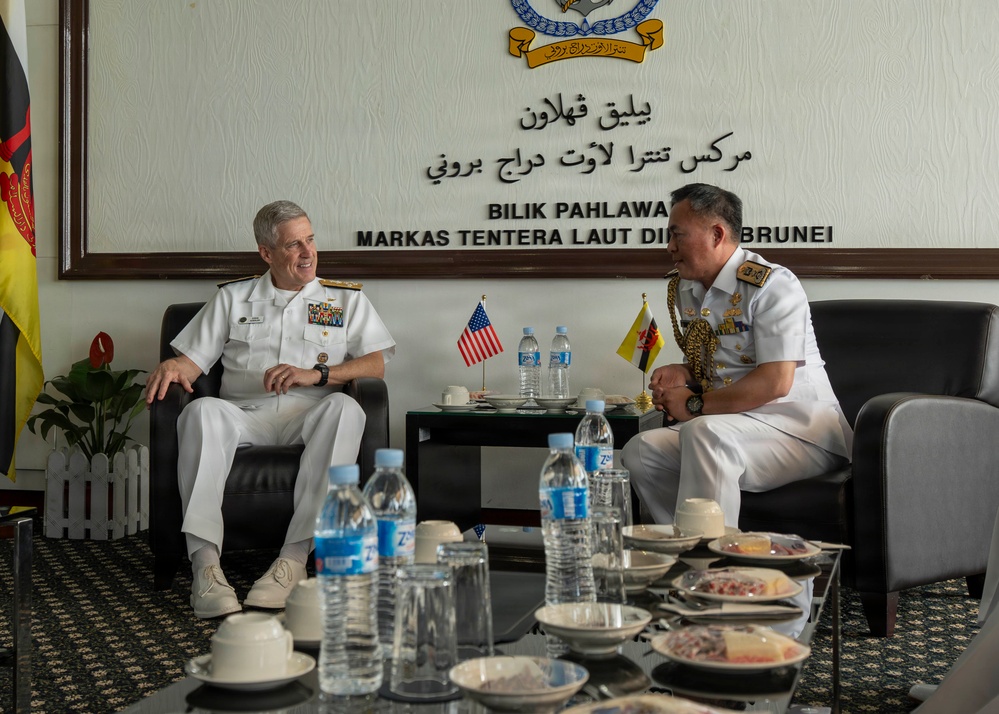 Two senior naval officers in white dress uniforms are engaged in a formal bilateral meeting at the Royal Brunei Navy headquarters (Markas Tentera Laut Diraja Brunei). One officer wears a US Navy uniform with multiple ribbons and insignia, while the other wears a Brunei Navy uniform with elaborate gold ceremonial decorations. Small US and Brunei national flags are displayed on the table between them, indicating an official diplomatic or military exchange.