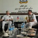 Two senior naval officers in white dress uniforms are engaged in a formal bilateral meeting at the Royal Brunei Navy headquarters (Markas Tentera Laut Diraja Brunei). One officer wears a US Navy uniform with multiple ribbons and insignia, while the other wears a Brunei Navy uniform with elaborate gold ceremonial decorations. Small US and Brunei national flags are displayed on the table between them, indicating an official diplomatic or military exchange.