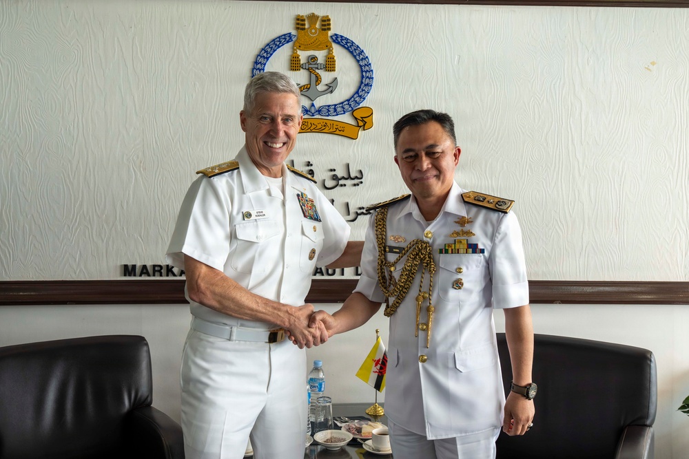 Two senior naval officers are shaking hands in what appears to be an official meeting room. The setting includes a naval emblem and Arabic/Malay text on the wall, suggesting this is a bilateral military engagement, likely between the United States Navy and the Royal Brunei Navy. Both officers are in formal white dress uniforms with rank insignia and decorations.