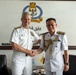 Two senior naval officers are shaking hands in what appears to be an official meeting room. The setting includes a naval emblem and Arabic/Malay text on the wall, suggesting this is a bilateral military engagement, likely between the United States Navy and the Royal Brunei Navy. Both officers are in formal white dress uniforms with rank insignia and decorations.